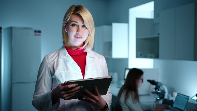 A blode female doctor in a laboratory checking something on the tablet before smiling at the camera. Another doctor is working on a microscope in the background. High quality 4k footage 
