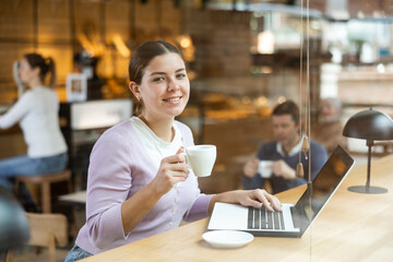 Fototapeta na wymiar Smiling young woman using laptop and drinking coffee in modern cafe