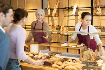 Two young old female bakery sellers helping with choice of delicious baked goods for customers behind counter in local bakeshop - obrazy, fototapety, plakaty