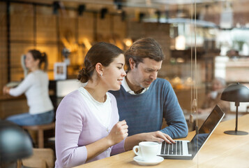 Middle-aged man and young woman sitting at table, watching video on laptop and enjoing cup of tea in coffee house
