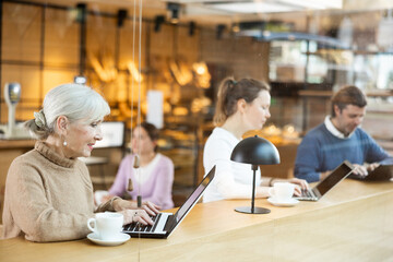 Smiling mature woman using laptop and drinking coffee in modern cafe
