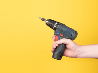 A hand holds a small cordless screwdriver. No face, yellow background, copy space.
