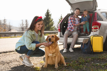 Happy woman petting dog, father and his daughter sitting in car trunk near road. Family traveling with pet