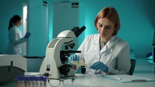 A veterinarian in a white lab coat prepearing a sample and looking at it through a microscope. There is another doctor in the background. High quality 4k footage 