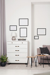 Empty frames hanging on white wall, chest of drawers and wooden tables indoors