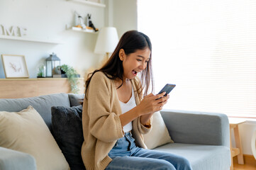 Happy young asian woman relax on comfortable couch at home texting messaging on smartphone, smiling girl use cellphone chatting, browse wireless internet on gadget, shopping online from home