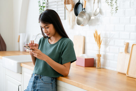 Happy young asian woman relaxing at home. Asia female standing at counter kitchen and using mobile smartphone
