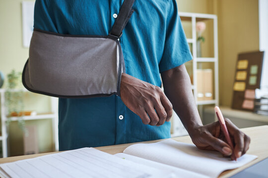 Close up of black man with arm sling working at standing desk in office and writing in notebook