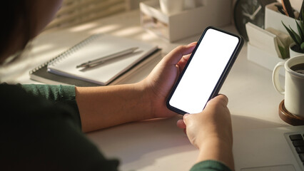 Over shoulder view of woman using mobile phone at working desk. Blank phone screen for create content and advertising.