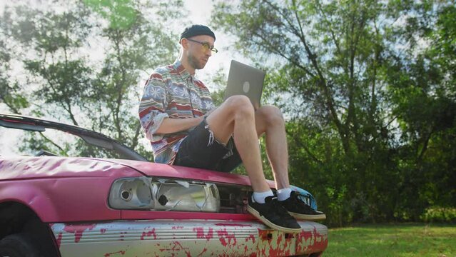Male freelancer remotely works via laptop sitting on hood of pink car outdoors. Young man works and focuses on computer screen. Stylish guy types on keyboard in park