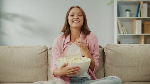 A Happy Young Woman Sitting on the Sofa, Eating Popcorn and Watching TV. Cheerful Woman Laughs Watching a Funny Movie. Movies as a Form of Relaxation and Psychological Relief. Online Cinemas.