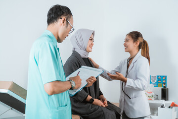 a female doctor smiling and touch the shoulder of the female patient that sitting on the bed next to the male nurse that standing with the medical check up paper at his hand