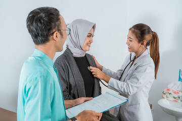 a beautiful doctor with long brown hair check up the female patient using the stethoscope with the male nurse standing beside the patient and bring the medical check up paper