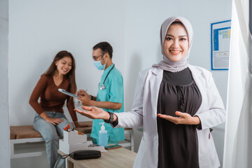 Obraz premium a beautiful doctor in doctor's coat standing with hand opened pointing on her side and smiling while the male nurse showing the medical check up result to the patient at the background