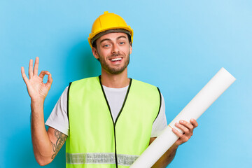 Young bearded man stands isolated over blue backdrop wearing yellow hard hat and holding in his...