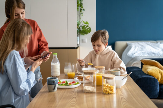 Mother and daughter using mobile phone at kitchen table while having breakfast together with family. Teen girl showing mom something on smartphone during mealtime. Cell phone usage and family bond - Powered by Adobe
