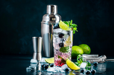 Naklejka na ściany i meble Blueberry Mojito cocktail drink with lime, white rum, soda, cane sugar, mint, and ice in glass on deep blue background. Summer refreshing beverage