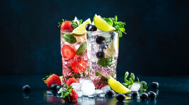 Strawberry and blueberry mojito cocktail drink with lime, white rum, soda, cane sugar, mint, and ice in highball glass on deep blue background. Summer refreshing beverage