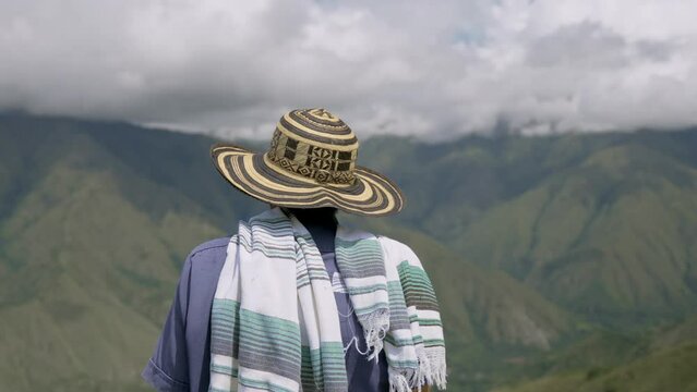 Colombian farmer with his hat looks at the majestic mountains in the distance, surrounded by lush green farmlands, symbolizing the vital role of nature in his livelihood in Llanos Orientale, Colombia.