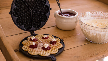An antique waffle iron with a freshly baked waffle in it decorated with raspberry jam and cream next to it there is a bowl with jam and one with cream