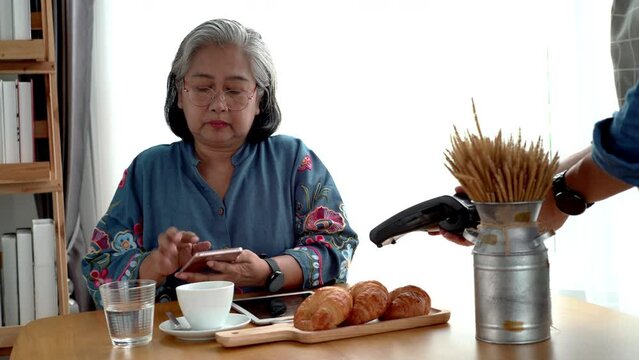 Active senior Asian woman working in a coffeeshop using a mobile device, smartwatch for NFC, QR code scanning, contactless payment to pay for coffee and snack. Active Senior concept.