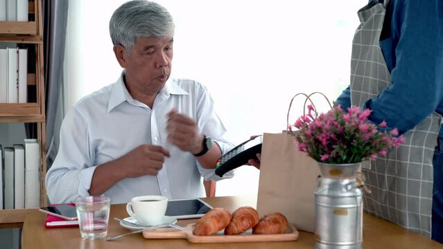 Active senior Asian man working in a coffeeshop using a mobile device, smartwatch for NFC, QR code scanning, contactless payment to pay for coffee and snack. Active Senior concept.