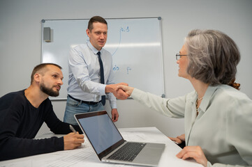 Obraz premium The boss makes a presentation to subordinates at the white board. Caucasian man shaking hands with middle aged woman. 