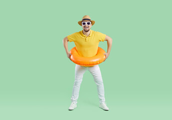 Happy man having fun on summer holiday. Full body length shot of cheerful funny young guy wearing yellow T shirt, hat, sunglasses and orange inflatable ring standing isolated on light green background