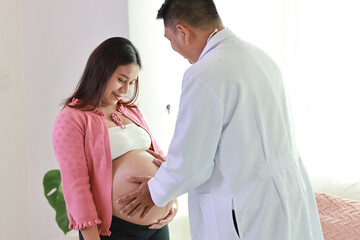 Happy smiling young asian pregnant female and male doctor sitting on sofa in living room at home with private treatment and consultant. Friendly doctor listening to baby’s heartbeat with stethoscope.