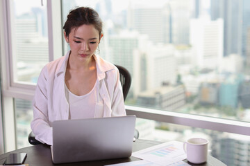 Portrait of confident and beautiful young asian businesswoman with happy emotion sitting and using computer in modern hotel with city blurred background. Success business and lifestyle concept