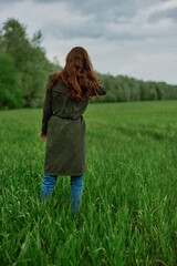 a woman with long red hair stands with her back to the camera in a field in tall green grass