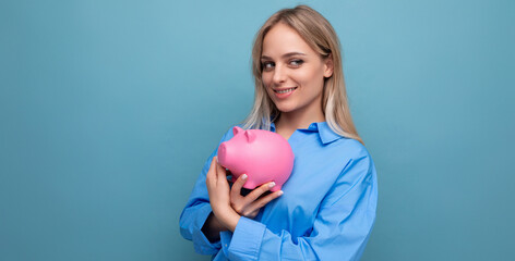 pensive blond young adult keeps savings in a piggy bank on a blue isolated background