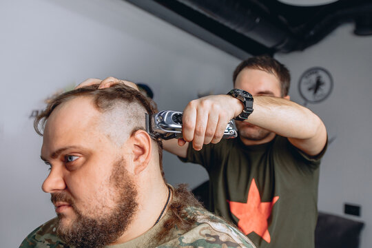A profile photo of an adult man with a brutal beard sitting in a barber shop with a black cape and barber's hands knocking down hair for a haircut.