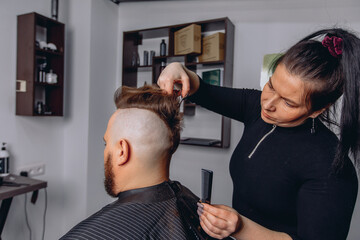 A master barber cuts his hair with scissors making a brutal mohawk for an adult bearded man. High-quality service in the barbershop. - obrazy, fototapety, plakaty