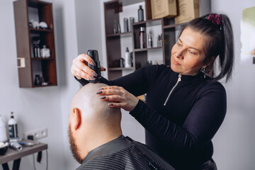 Girl hairdresser shaves an adult man with beard baldly using electric razor in barbershop holding it in her hand. Provision of hairdressing services.