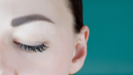 Close-up of closed eye with extreme long eyelashes, black eyeliner, healthy skin, perfect eyebrow of unrecognizable young woman on green background. Macro. Lash extension, beauty procedures, make-up.