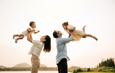 A cheerful Asian family enjoys a day out in nature, as the father and mother throw their toddler up into the sunny sky. The happy child enjoys the playful freedom of flying, captured in a photograph