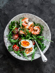 Breakfast, snack  bowl - arugula, cherry tomatoes salad with boiled egg and fried shrimp on with sesame olive oil dressing a dark background, top view