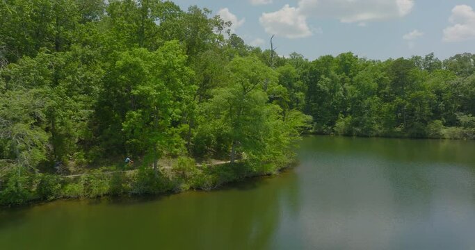 Aerial: Friends Riding Bicycles In Green Forest, Drone Flying Forward Over Rippled River On Sunny Day - Tuscaloosa, Alabama