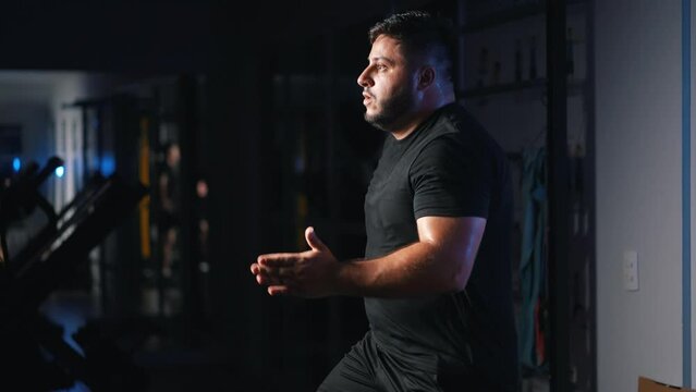 Energetic overweight man running in place as a cardio exercise for daily workout in a dark gym training hard for competition in slow motion