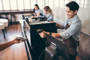 Happy Asia man waiter in apron with card reader and customer with smartphone paying at bar of coffee shop