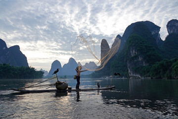 An old man stands on a bamboo raft in front of the Guilin landscape background and casts a net for...