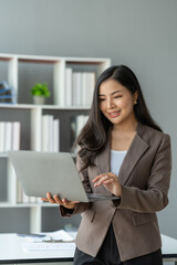 Asian businesswoman smiling confidently successful entrepreneur in the financial business professional company executive Wearing suit standing holding laptop searching business information in office