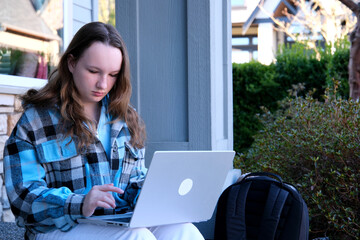 teenage girl with a laptop sitting on the porch of house. She watching the news doing homework...