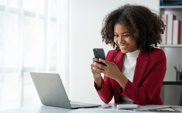 African American business woman holding a mobile phone to read financial business news Online marketing on mobile apps to process emails in meetings sitting at laptop desk in office.