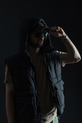 Cool fashionable man with sunglasses in trendy black clothes puts on a cap in the dark. Fashion guy on a black background