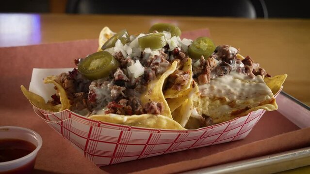 Paper boat filled with barbecue brisket nachos, tortilla chips covered with chopped smoked brisket queso pickled jalapenos chopped onions, close up slider 4K
