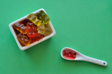 multi-colored tasty gummy bears on a small plate and a small spoon on a green background
