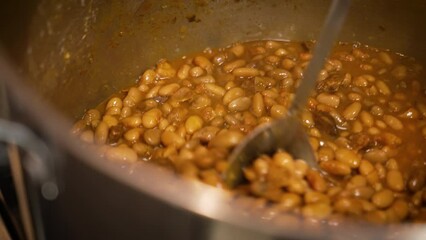 Large spoon stirs huge pot filled with barbecue pinto beans, slow motion close up 4K