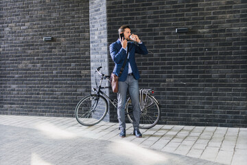 Caucasian men in suit standing in front of his bicycle, he using his phone and drinking morning...